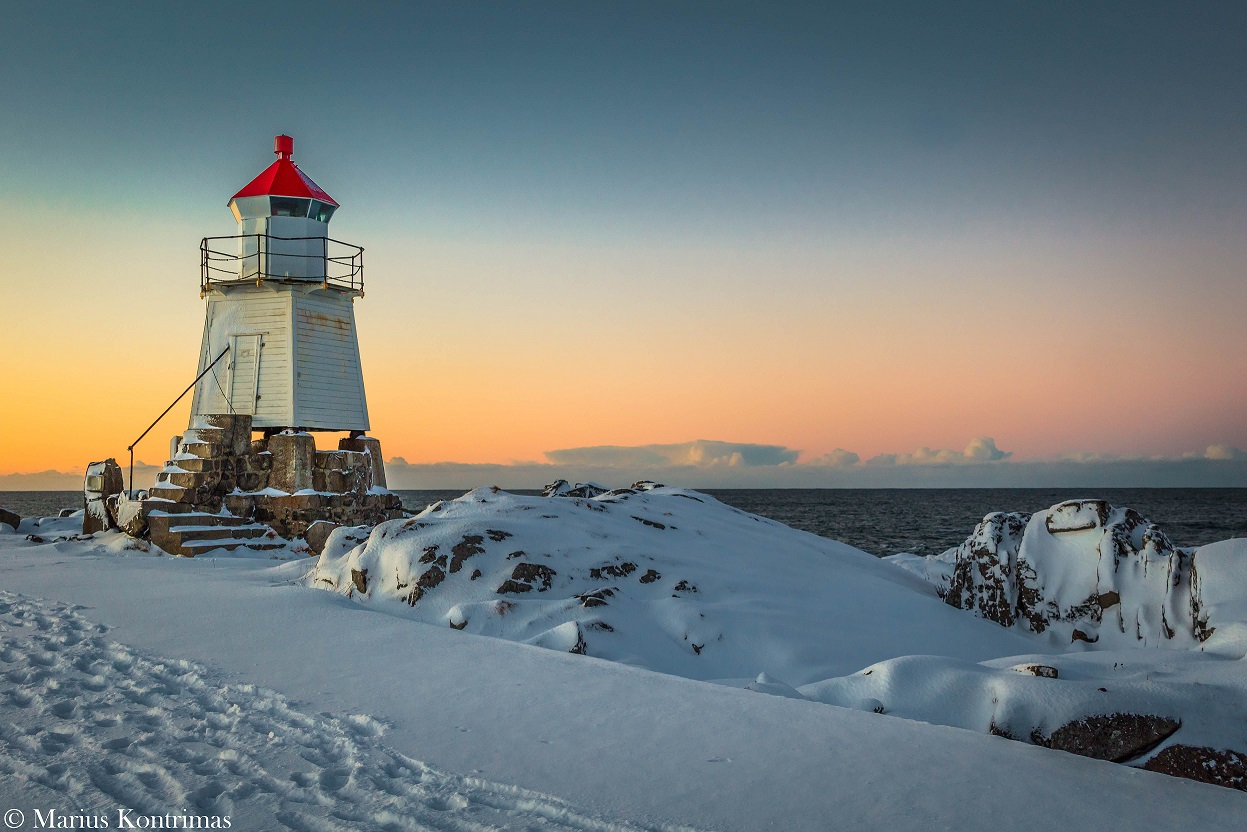 A red-roofed lighthouse on a snow-covered shore at sunset above the Arctic sea